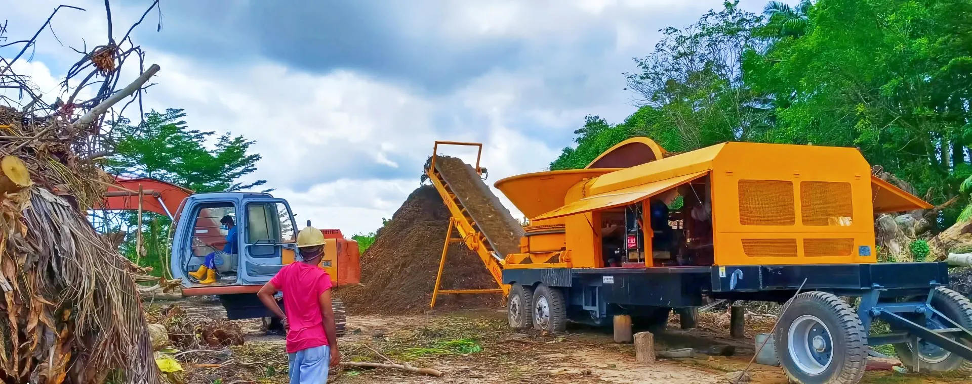 WeiDong 3600-series wheeled tub grinder grinding tree and palm residue fed by an excavator, with the discharge conveyor building a stockpile