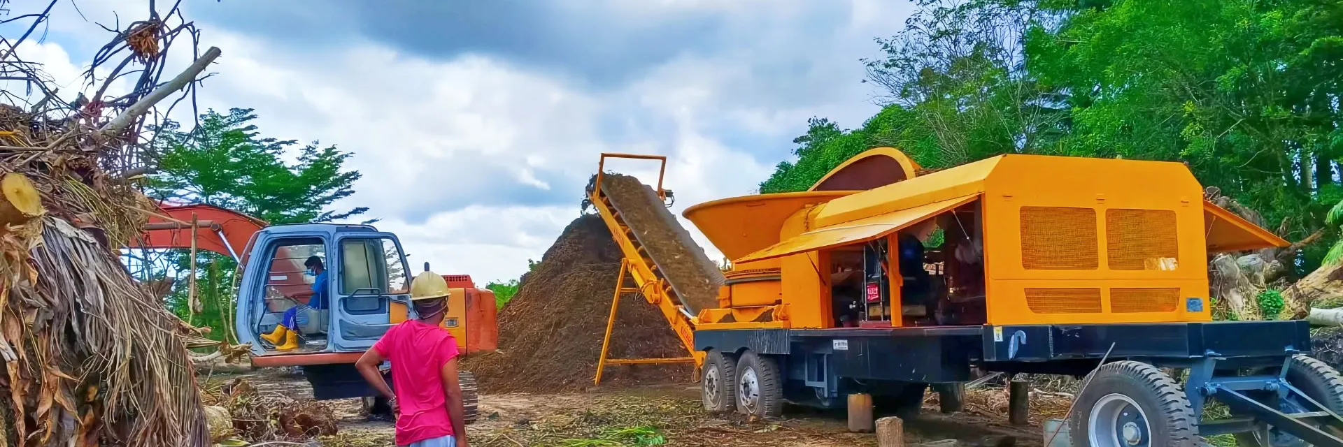 WeiDong 3600-series wheeled tub grinder grinding tree and palm residue fed by an excavator, with the discharge conveyor building a stockpile